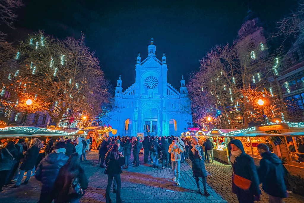 Brussels Christmas Market at night in front of Sainte-Catherine Church, glowing with blue lighting and surrounded by festive wooden stalls and twinkling tree lights.