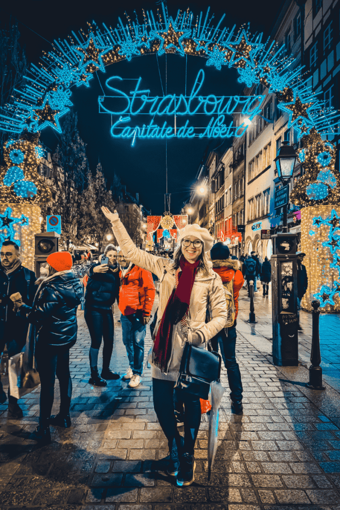 Kate stands under the glowing blue “Strasbourg Capitale de Noël” arch, smiling and pointing up at the festive entrance to the Christmas market.