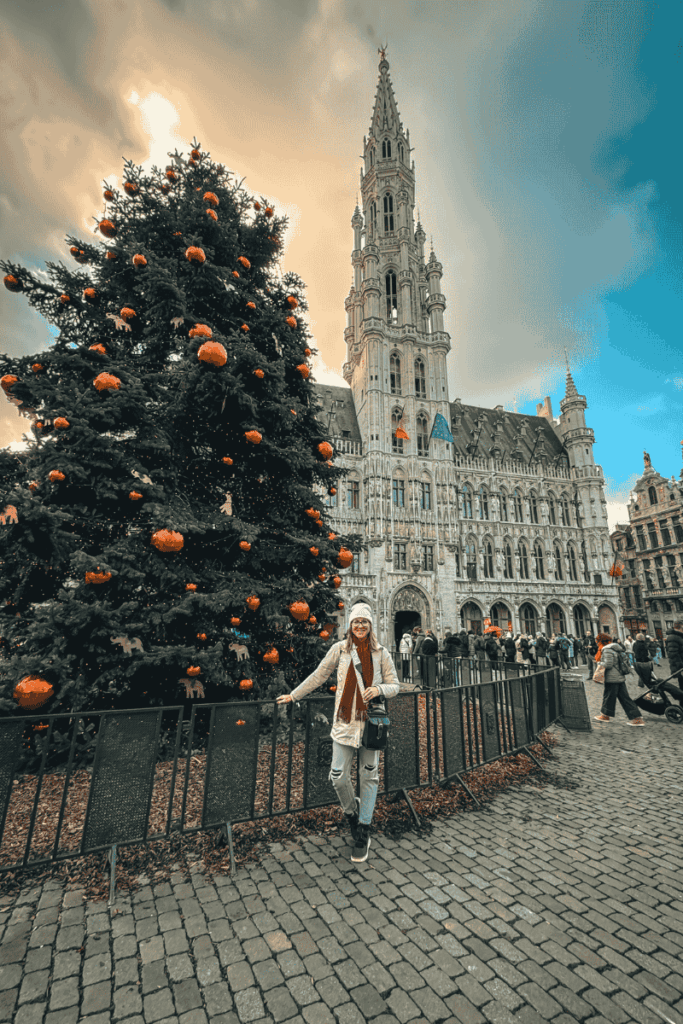 Kate poses beside a Christmas tree decorated with orange ornaments in a grand square with a Gothic-style building in the background.