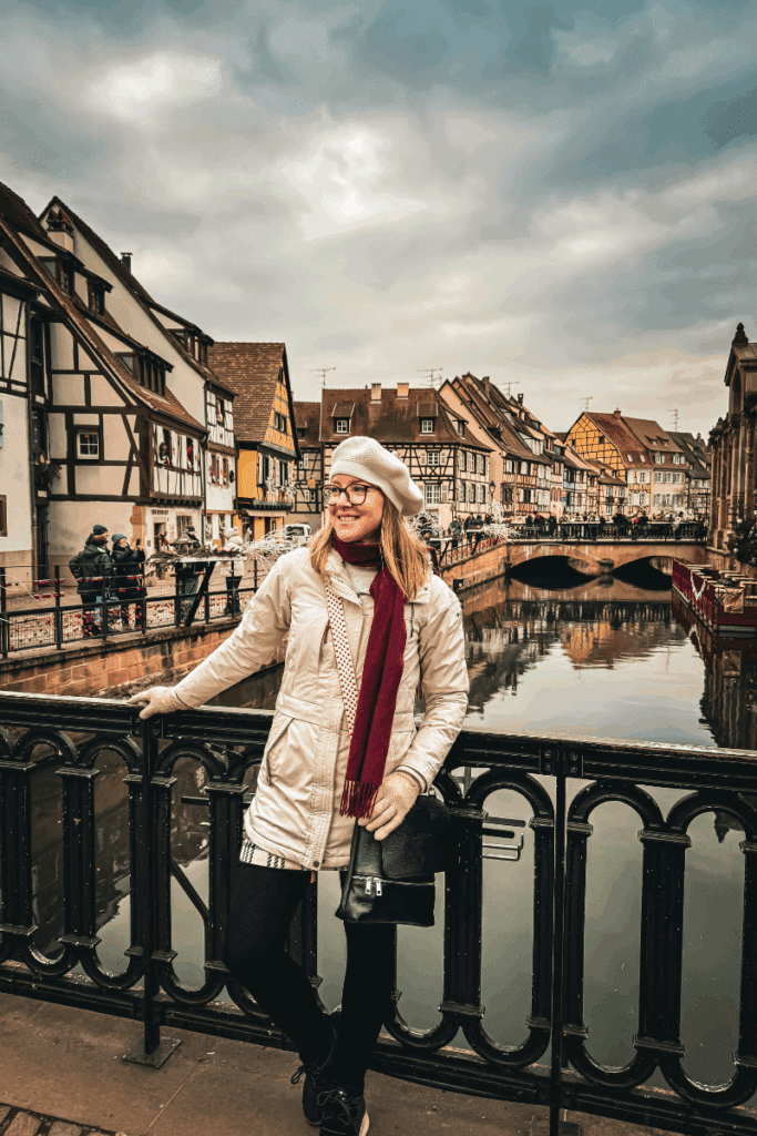 Kate leans against a black railing on a bridge overlooking a canal lined with charming half-timbered houses in a European village.