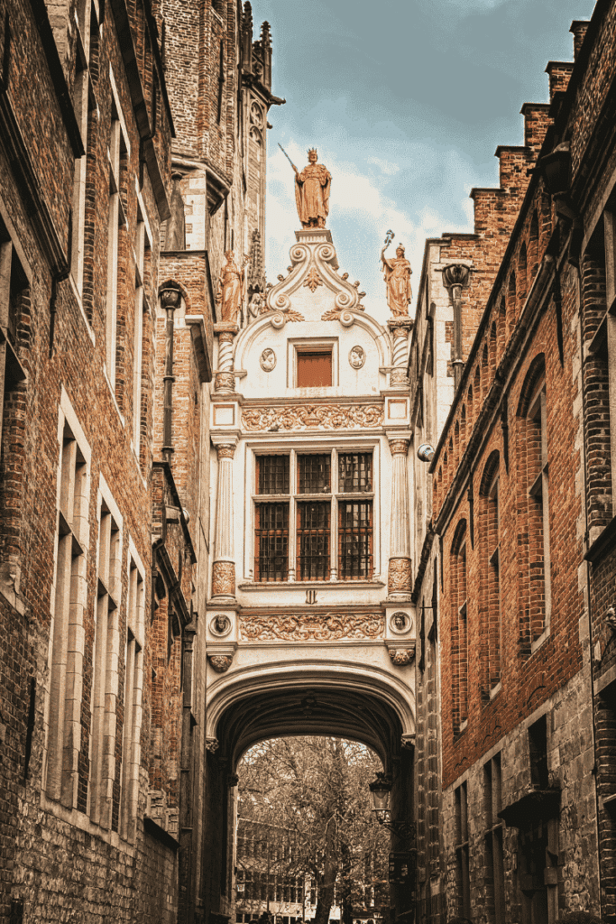 An ornate white and gold arched passageway adorned with statues connects two brick buildings along a narrow cobblestone alley.