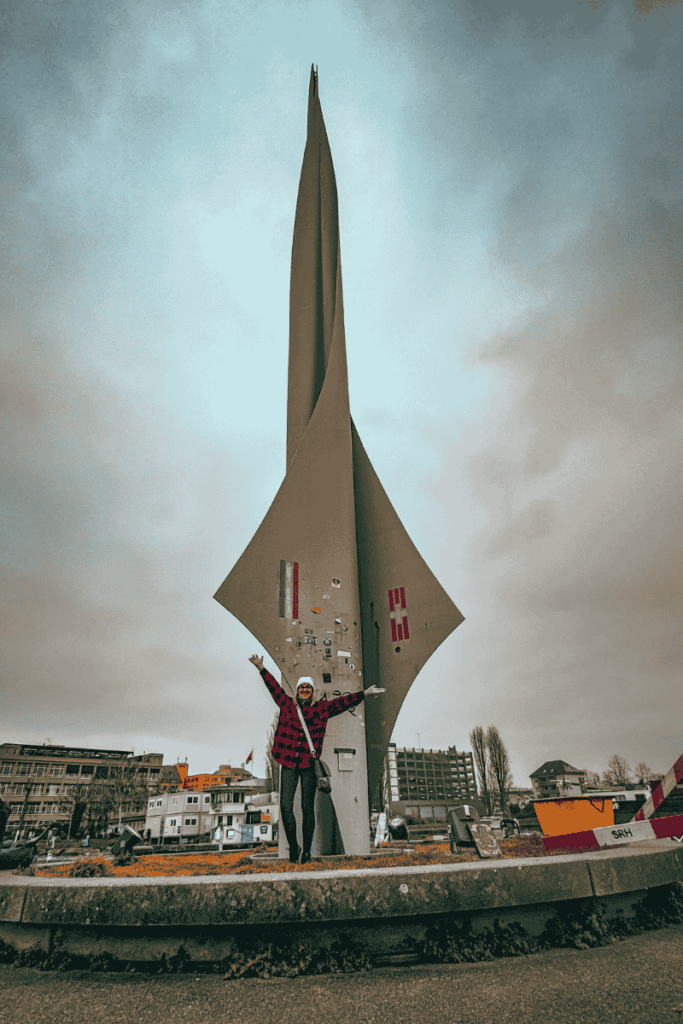 Kate poses with arms raised in front of a tall, futuristic-looking concrete sculpture covered in stickers at a roundabout.