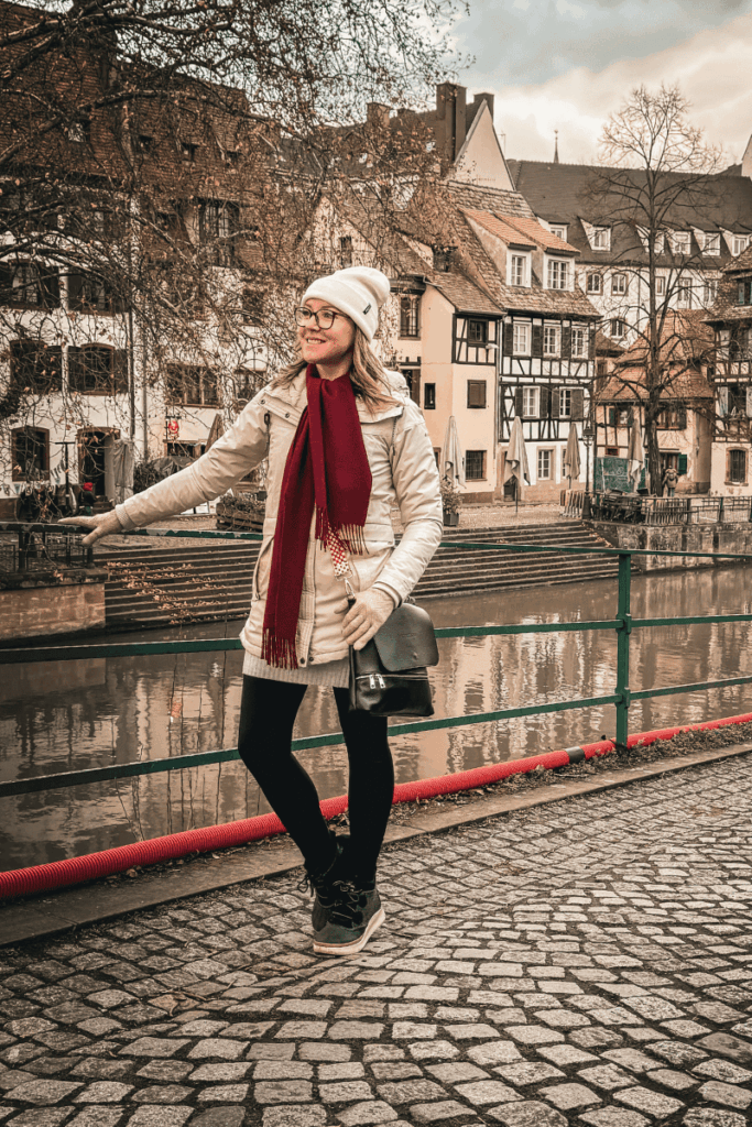 Kate stands along a riverside path lined with half-timbered houses, smiling and holding onto a railing in a quaint European town.