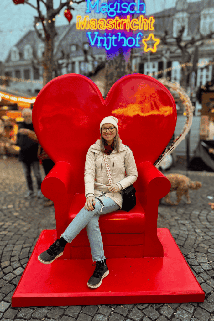Kate sits on a giant red heart-shaped chair under glowing “Magisch Maastricht Vrijthof” lights at a Christmas market.