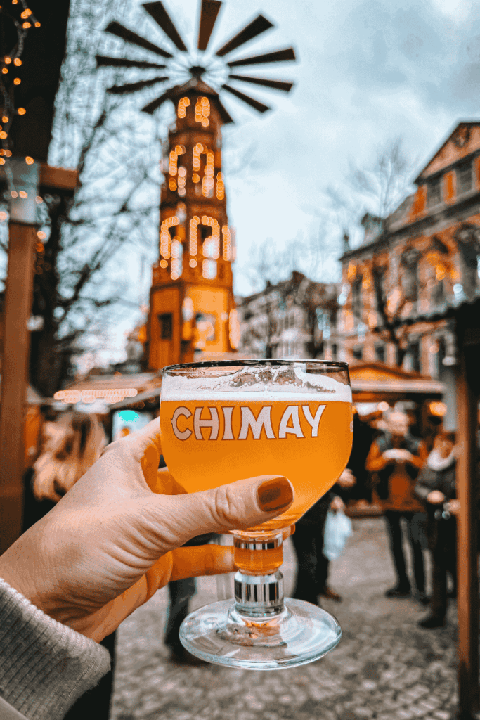 A close-up of a hand holding a glass of Chimay beer with a glowing wooden Christmas pyramid blurred in the background.