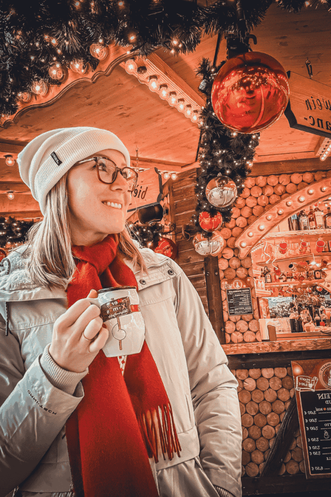 Kate smiles while holding a festive mug inside a cozy Christmas market stall decorated with garlands and oversized red ornaments.