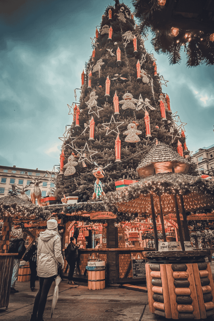 Kate stands in front of an enormous Christmas tree adorned with red candle lights, stars, and gingerbread figures at a lively holiday market.