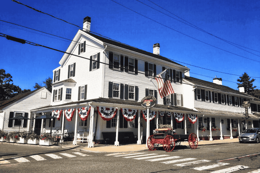 The historic Griswold Inn in Essex, Connecticut features classic white siding, black shutters, patriotic bunting, and a red antique wagon parked out front under a clear blue sky.