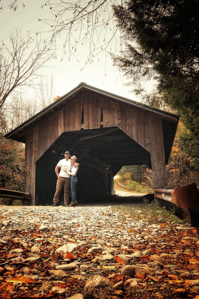 Kate and her husband stand arm in arm on a rocky path in front of a rustic wooden covered bridge surrounded by early autumn trees along Vermont's scenic Route 100.
