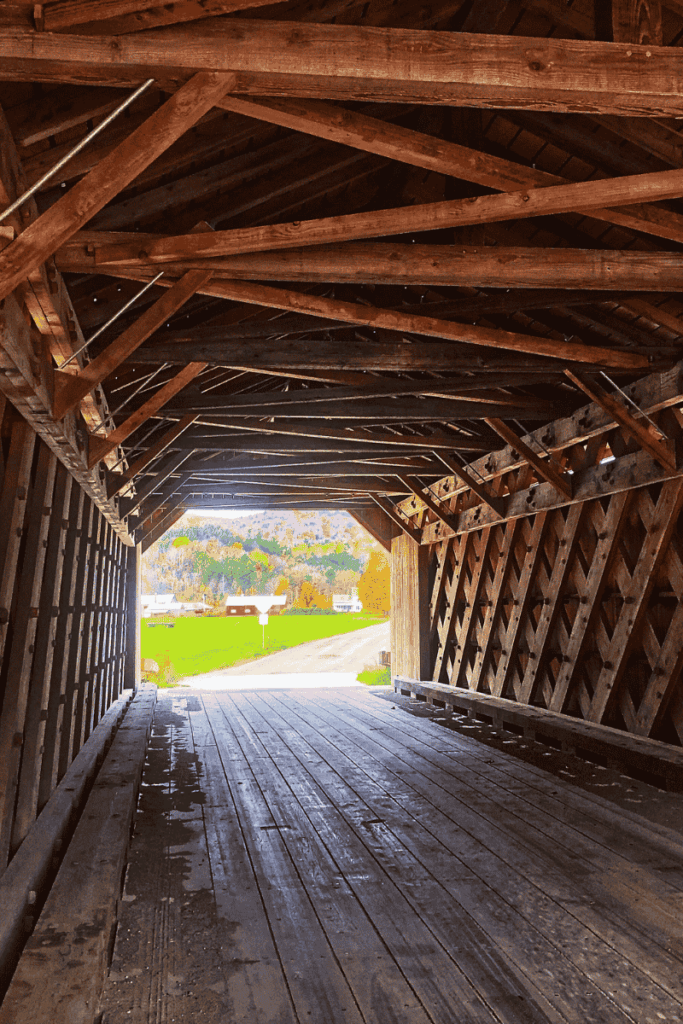 View from inside a wooden covered bridge looking out onto a rural Vermont road, with bright green fields and colorful autumn hills in the distance.