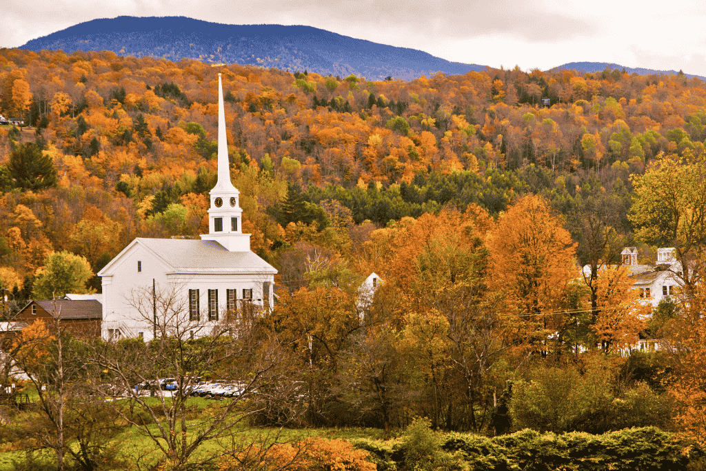 A white church with a tall steeple stands nestled in the heart of Stowe, Vermont, surrounded by rolling hills blanketed in vibrant fall foliage.