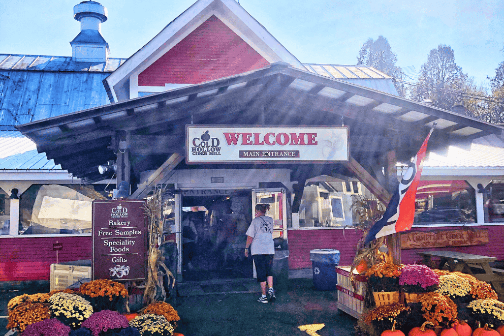 A visitor walks into the welcoming entrance of Cold Hollow Cider Mill in Vermont, surrounded by colorful mums, pumpkins, and signs advertising bakery treats and specialty foods.