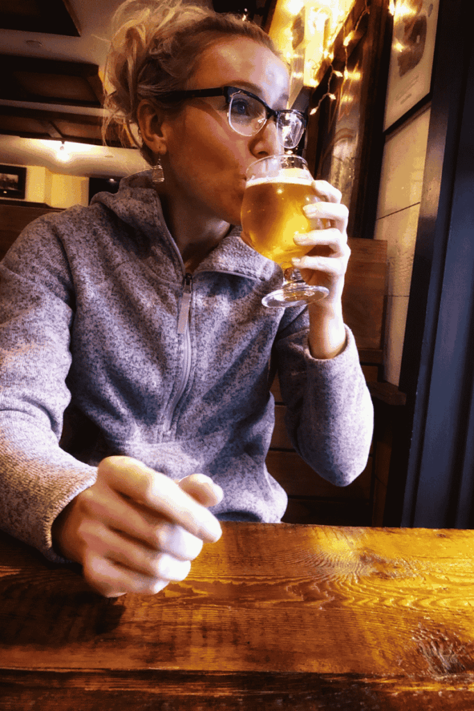 Kate sips a glass of golden craft beer in a cozy, warmly lit restaurant.