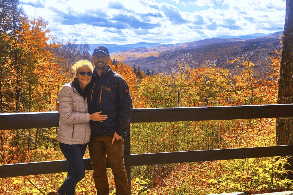 Kate and her husband smile together on a scenic overlook with a sweeping view of colorful fall foliage and distant New England mountains.