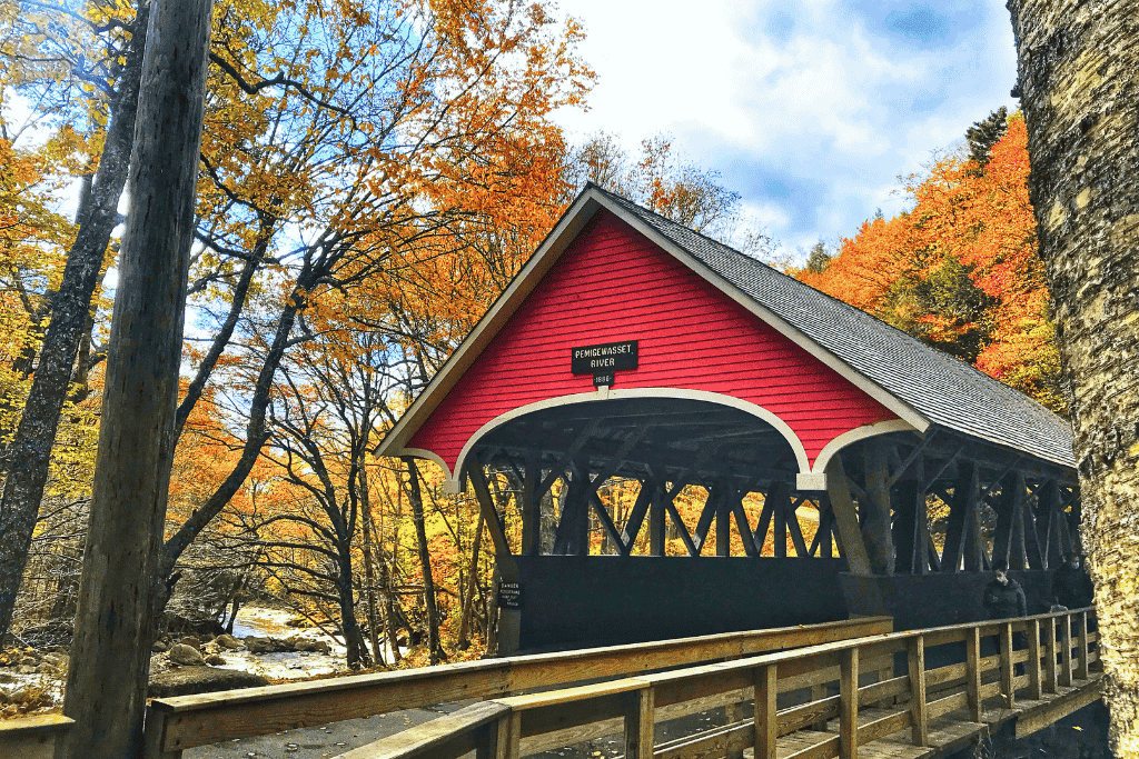 The red Pemi Bridge crosses the Pemigewasset River in a blaze of fall color, surrounded by golden foliage and clear mountain light in New Hampshire.