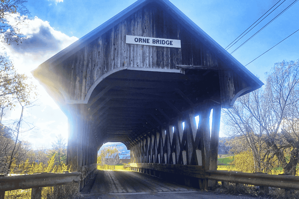 Sunlight filters through the weathered wooden structure of Orne Bridge, framing a quiet country road and distant hills beyond.