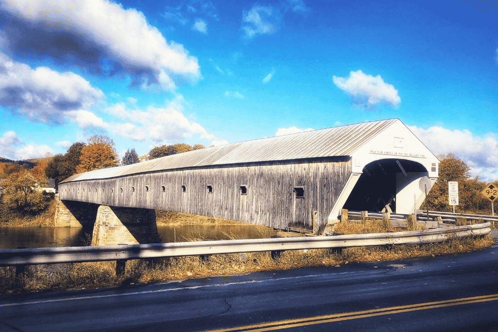 The Cornish-Windsor Covered Bridge stretches across the Connecticut River under a bright blue sky, its long wooden frame flanked by autumn trees and road signs marking the Vermont-New Hampshire border.