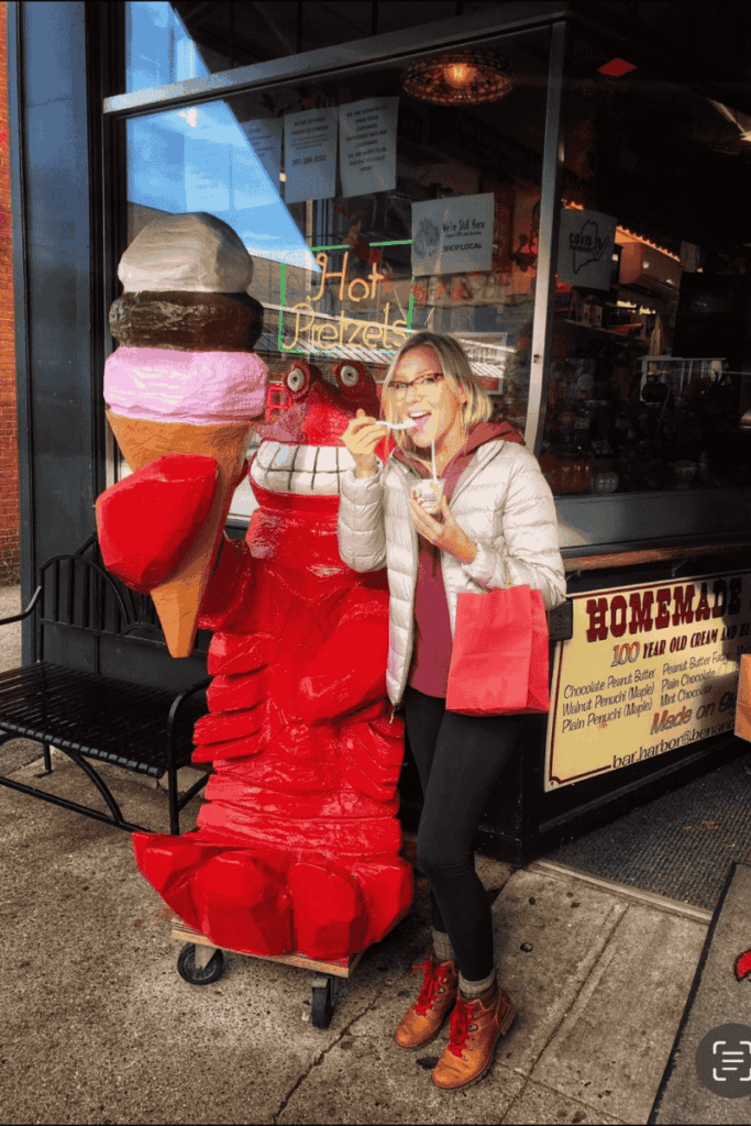 Kate smiles while eating ice cream next to a quirky red lobster statue holding a giant cone outside a homemade ice cream shop in Bar Harbor, Maine.