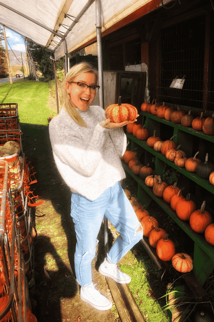 Kate beams while holding a striped mini pumpkin at an outdoor roadside farm stand lined with shelves of gourds and squashes.