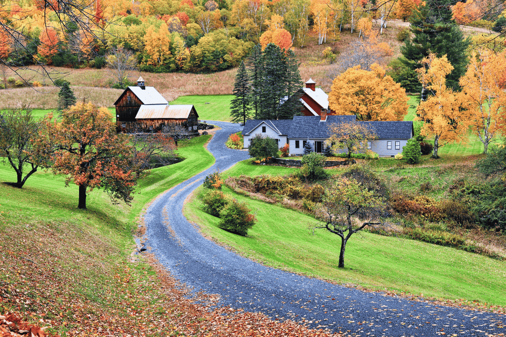 A winding gravel road leads to the picturesque Sleepy Hollow Farm in Vermont, surrounded by rolling hills, rustic barns, and a vibrant patchwork of fall foliage.