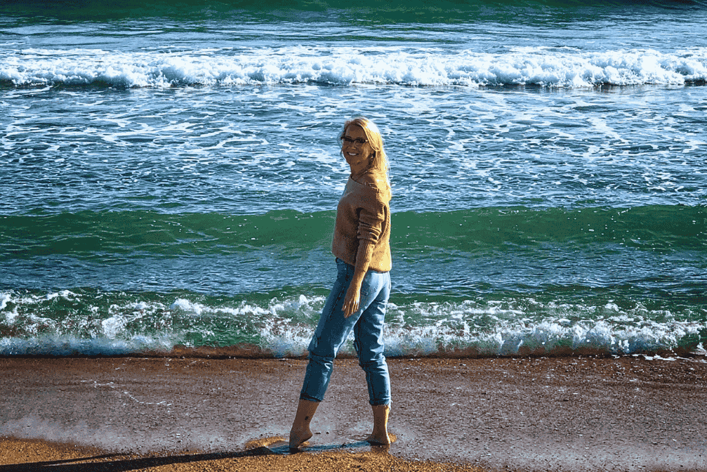 Kate smiles barefoot at the edge of the surf on a Newport, Rhode Island beach, with green-blue waves rolling in behind her.
