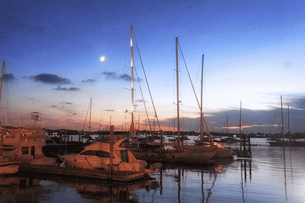 Sailboats and yachts rest peacefully in a Newport, Rhode Island marina at dusk, with a glowing moon and soft sunset colors reflecting off the calm water.