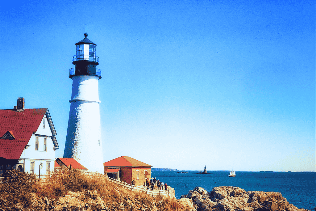 A classic white lighthouse with a red-roofed keeper’s house stands on a rocky Maine coastline under a clear blue sky, with a sailboat and distant lighthouse visible on the horizon.