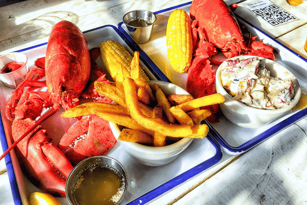 Two trays of whole steamed lobsters come with corn on the cob, French fries, and creamy potato salad, ready for a classic Maine seafood feast.