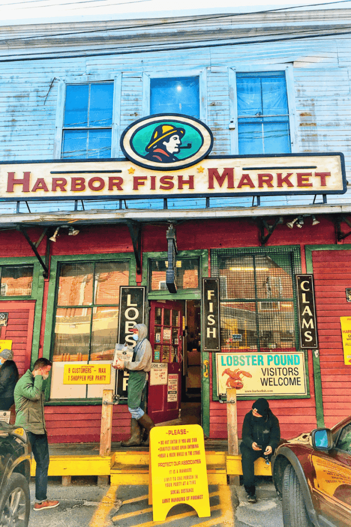 The colorful facade of Harbor Fish Market in Portland, Maine features bold signage and customers waiting outside under pandemic-era shopping guidelines.
