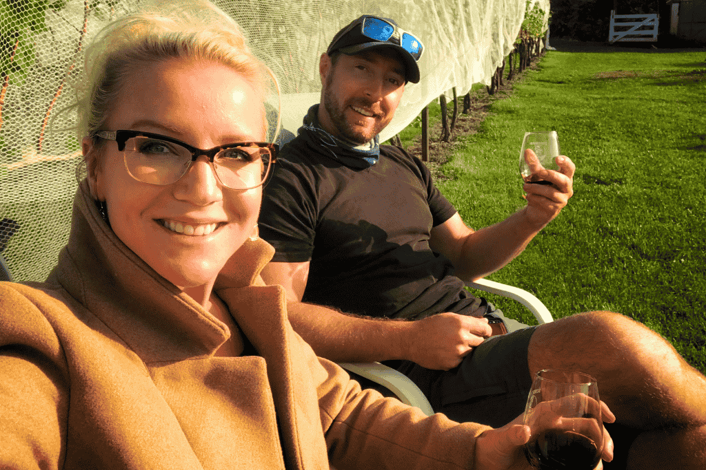 Kate and her husband relax with glasses of red wine on vineyard chairs, smiling in the golden late afternoon sun.