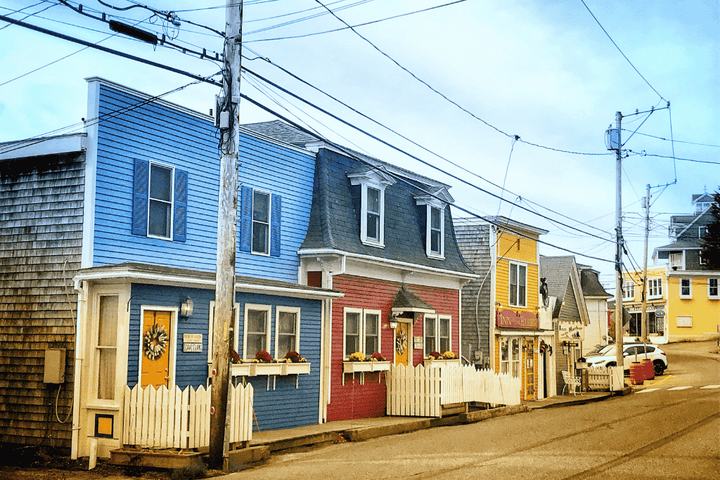 A row of colorful storefronts and cottages with white picket fences lines a quiet sloped street in the coastal village of Stonington, Maine.