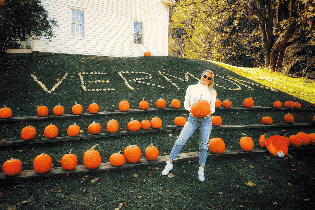 Kate stands smiling in front of a hillside lined with pumpkins and a white-gourd arrangement spelling “VERMONT,” holding a large pumpkin on a sunny fall day.