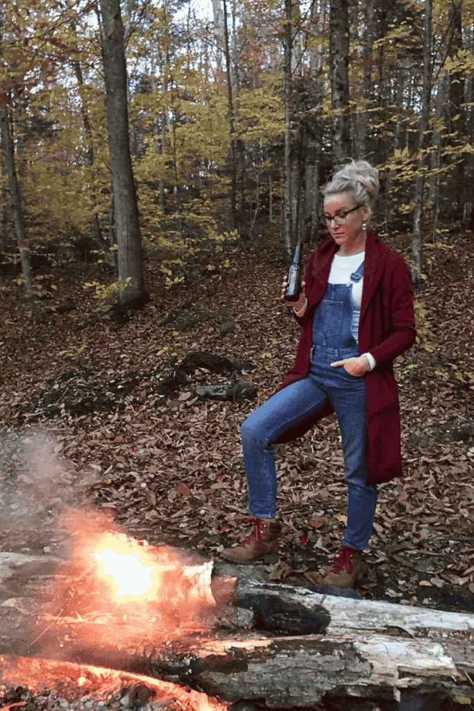 Kate stands by a crackling campfire in the woods, holding a bottle of beer and dressed warmly in overalls, boots, and a long cardigan on a crisp autumn evening.