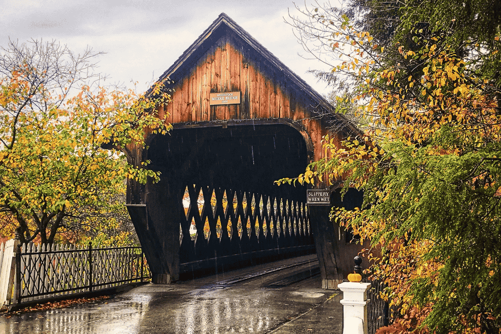 The Woodstock Middle Bridge in Vermont stands wet from rain, framed by golden autumn leaves and a sign that reads “Slippery When Wet,” capturing the charm of a classic New England fall day.
