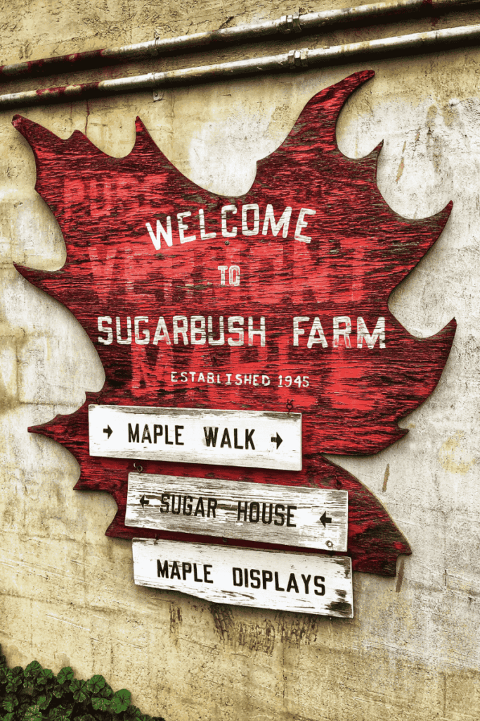 A large red maple leaf-shaped sign welcomes visitors to Sugarbush Farm in Vermont, with weathered wooden arrows pointing to the Maple Walk, Sugar House, and Maple Displays.