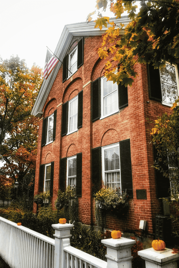 A stately red brick home in Woodstock, Vermont features black shutters, window boxes, and small pumpkins lining a white picket fence, all framed by autumn foliage and an American flag.