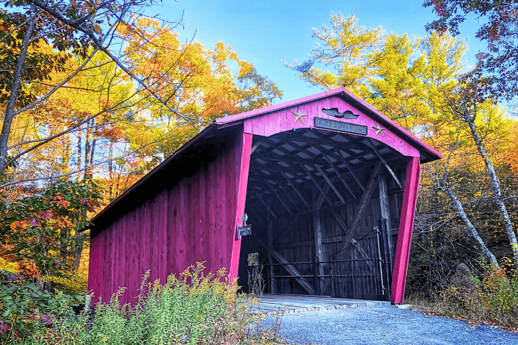 The bright red Tannery Bridge in Vermont stands out against a backdrop of colorful fall foliage, with golden trees framing its rustic wooden structure under a clear blue sky.