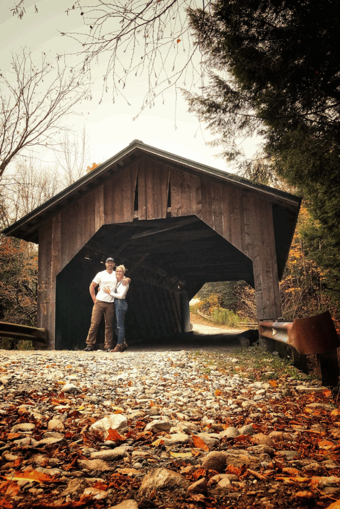 Kate and her husband stand arm in arm at the entrance of a rustic covered bridge surrounded by fall leaves and trees.