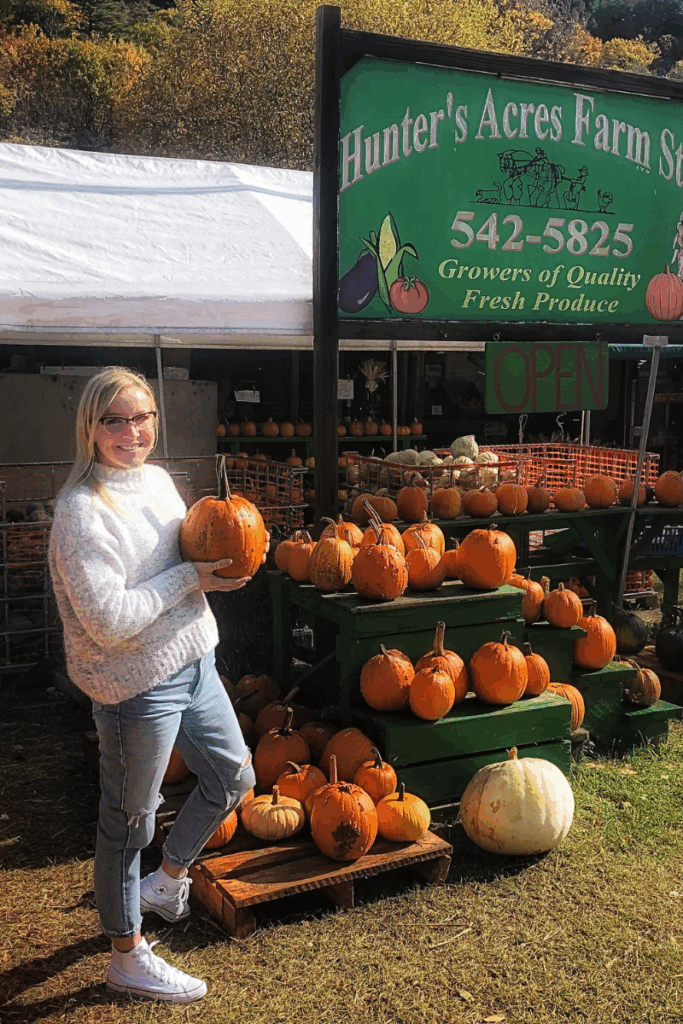 Kate smiles while holding a pumpkin at Hunter’s Acres Farm Stand, surrounded by stacked pumpkins under a green sign.