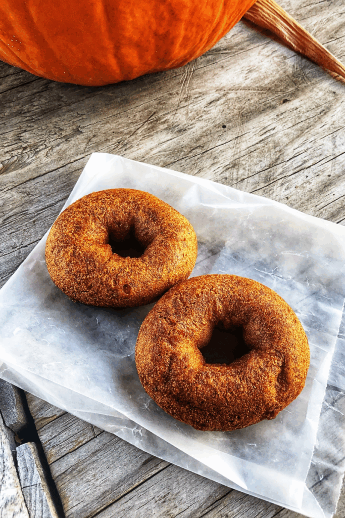 Two fresh apple cider donuts rest on wax paper beside a pumpkin on a rustic wooden table.