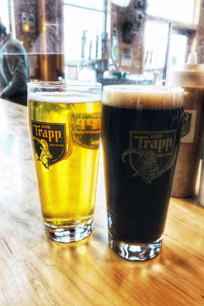 Two beers from von Trapp Brewing—a golden lager and a dark stout—sit on a wooden bar, with their logos clearly visible in a cozy Vermont taproom.