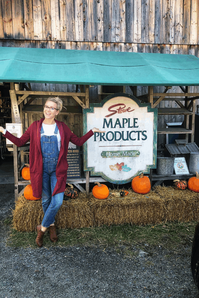 Kate poses cheerfully in front of a “Stowe Maple Products” sign with pumpkins and hay bales outside a rustic Vermont building.