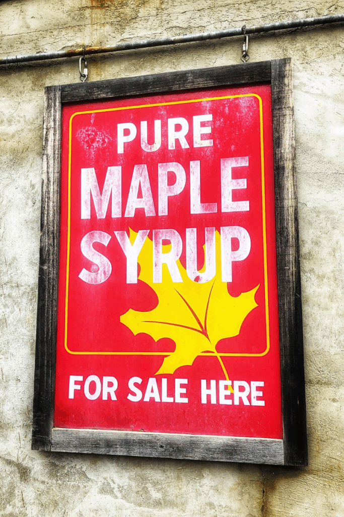 A bright red sign with a yellow maple leaf reads “Pure Maple Syrup For Sale Here” against a concrete wall.