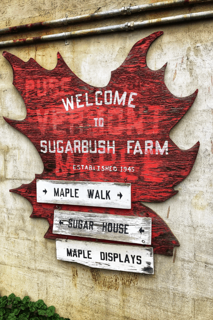 A large red wooden maple leaf sign welcomes visitors to Sugarbush Farm, pointing to the maple walk, sugar house, and displays.