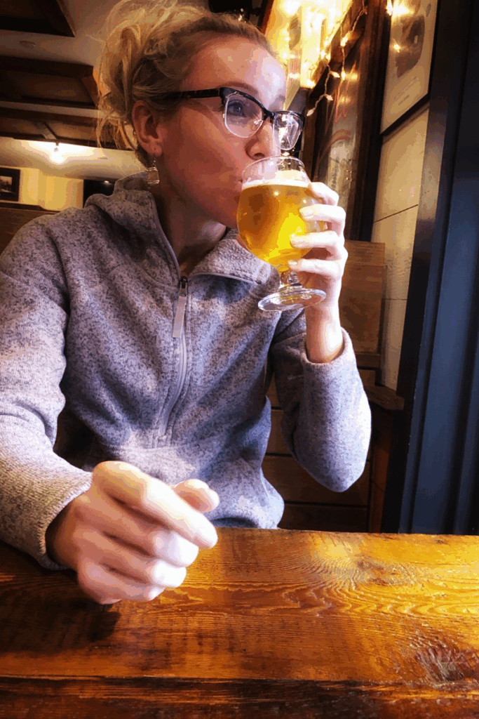 Kate sips a golden beer from a stemmed glass while sitting at a wooden table inside Worthy Kitchen in Vermont.