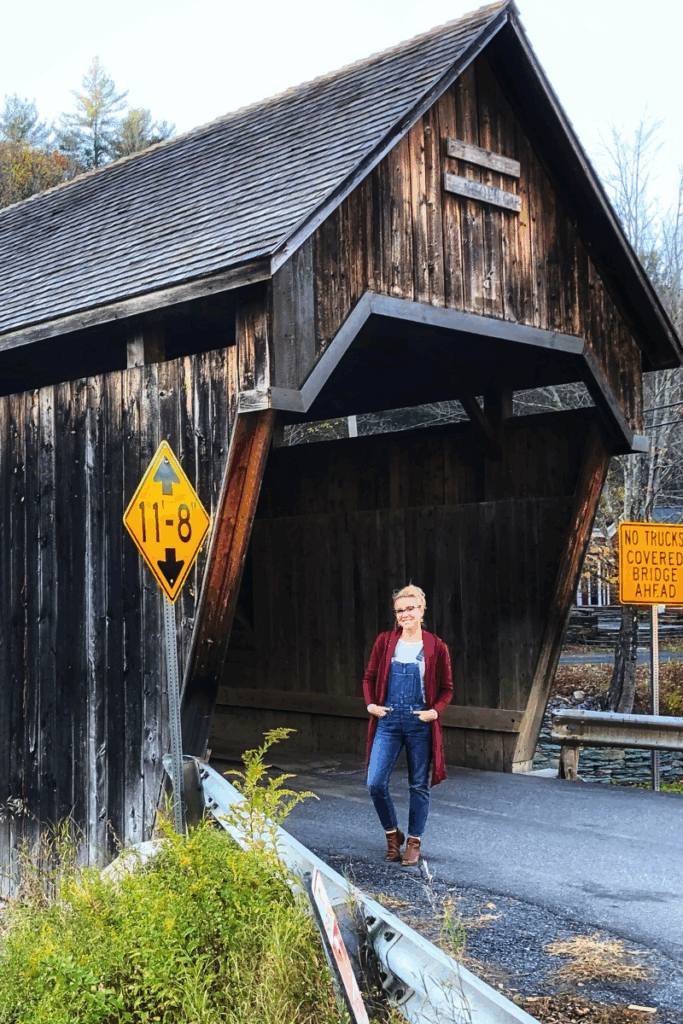 Kate stands smiling in front of a tall wooden covered bridge in Vermont, framed by road signs and trees in early fall.