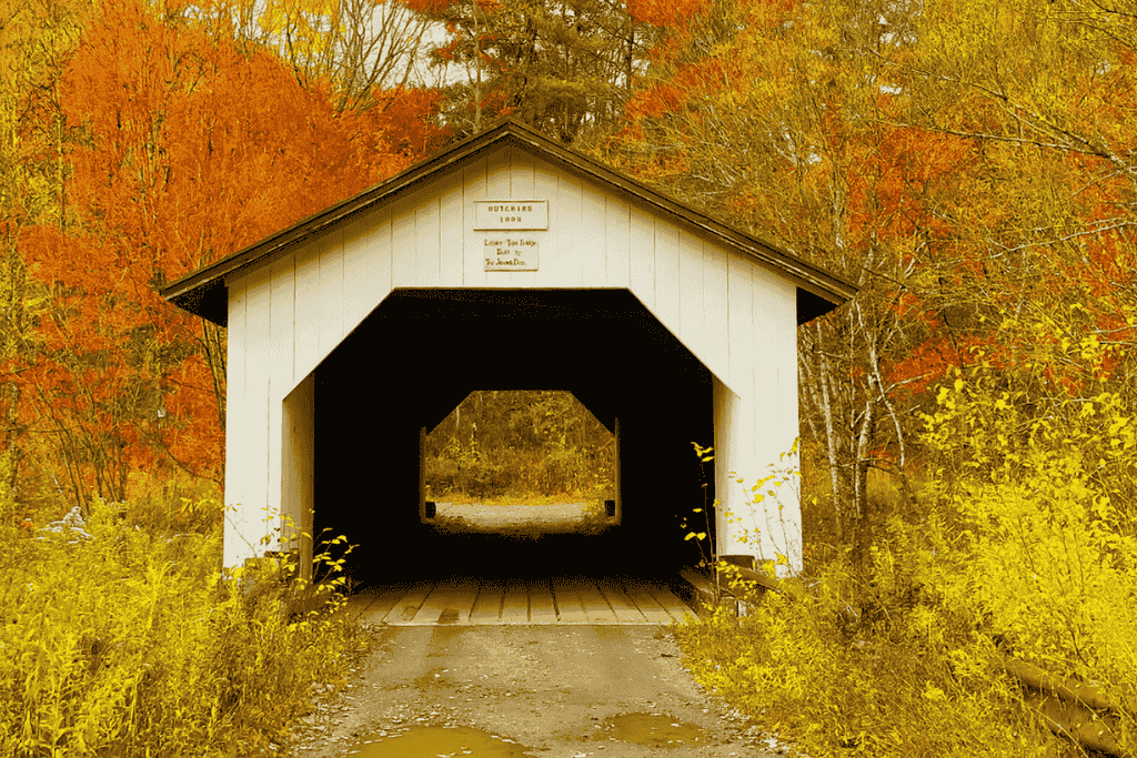 The white Hutchins Covered Bridge in Vermont stands framed by vibrant fall foliage in shades of yellow, orange, and red, capturing a classic New England autumn scene.