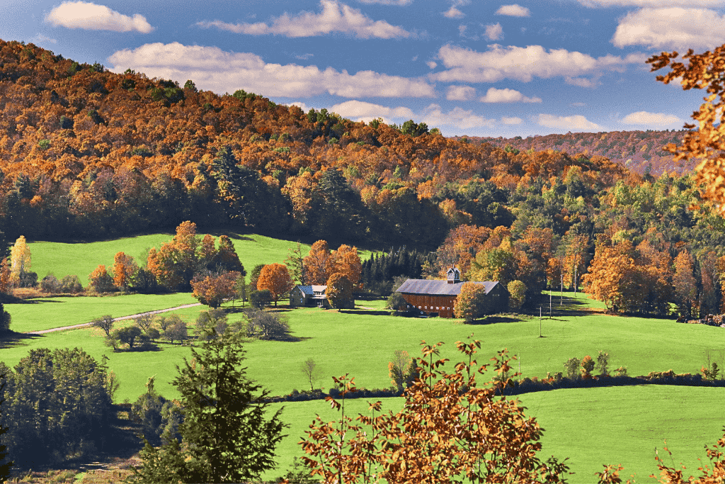 A scenic view along Vermont’s Highway 100 shows a peaceful farmstead nestled among rolling green fields and forested hills bursting with vibrant fall foliage.
