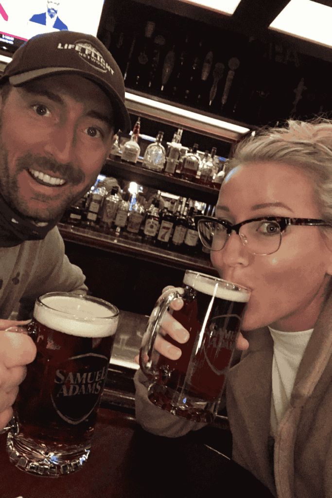 Kate and her husband enjoying pints of Samuel Adams beer at a cozy Boston bar, smiling with a fully stocked bar in the background.