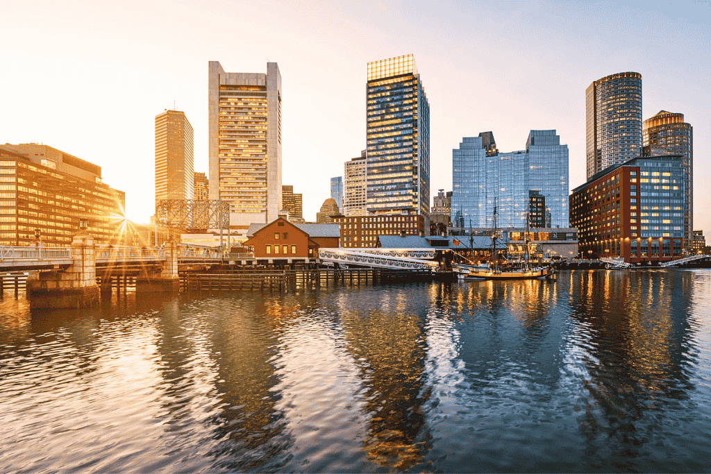 Boston Harbor at sunset with tall modern buildings reflecting in the water and a historic ship docked near the Boston Tea Party Ships & Museum. 